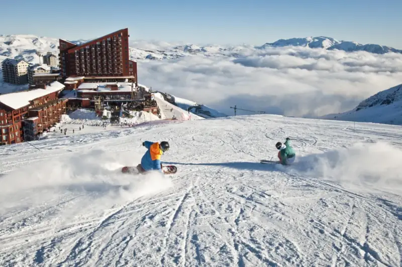 Duas pessoas descendo de snowboard na pista do Valle Nevado, com o hotel à esquerda e um mar de nuvens ao fundo