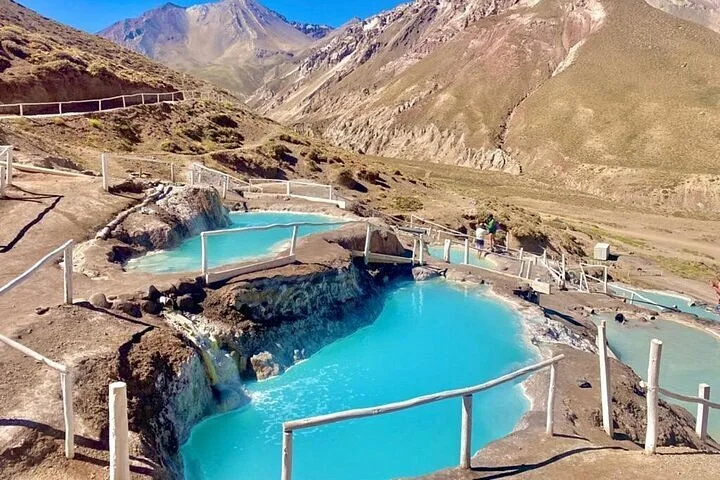 Piscinas de água turquesa das Termas de Colina em degraus na encosta, cercadas por corrimãos de madeira, com a montanha seca ao fundo