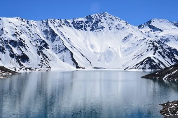 Lago azul do Embalse El Yeso espelhando as montanhas cobertas de neve em volta, sob céu limpo