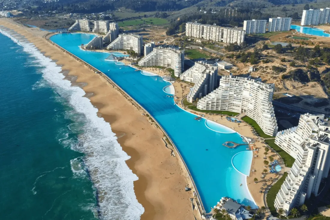 Vista de cima da piscina gigante de Algarrobo, de água azul, correndo ao lado da praia e do mar aberto, com os prédios do condomínio à direita