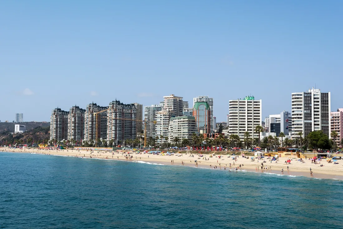 Praia de Viña del Mar com mar azul turquesa, faixa larga de areia e a fileira de prédios e palmeiras da orla